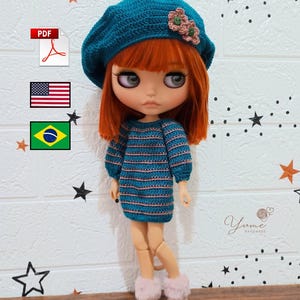 Pode incluir: Uma boneca vestindo uma boina de crochê azul-petróleo com detalhes de flores rosa, um vestido listrado combinando e botas rosa com acabamento de pele. A boneca tem cabelo ruivo e está posada contra uma parede de tijolos brancos com estrelas.