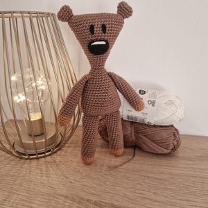 Può includere: Bambola orso all'uncinetto marrone fatta a mano con testa triangolare, piccole orecchie e bocca nera. L'orso ha braccia e gambe piccole con punte color cuoio. Una lampada in filo d'oro e gomitoli di filo sullo sfondo.