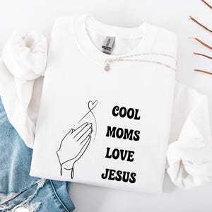 May include: White sweatshirt with the text "COOL MOMS LOVE JESUS" in black. The design includes a line drawing of praying hands and a heart. A silver necklace with a pendant is draped across the sweatshirt.