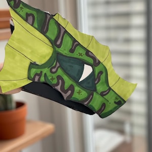 May include: A handmade dragon mask in lime green and dark green. The mask has a detailed eye and camouflage patterns. The mask is made of paper and has a black base.