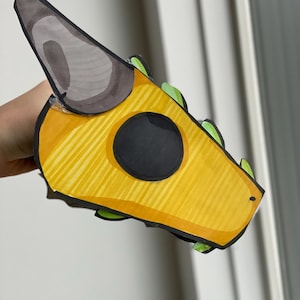 May include: A handmade, yellow dragon head mask with black and green accents. The mask features a large black eye and a grey horn. The dragon head is made of paper and has a striped pattern.