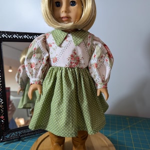 May include: A doll with blonde hair and blue eyes wearing a green and white floral dress with a green polka dot skirt. The doll is wearing brown boots and is displayed on a wooden stand.