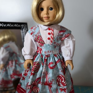 May include: A doll wearing a Christmas-themed dress. The dress features a light blue background with candy canes, lollipops, and the words "Jolly" and "Have". The dress has a white collar and sleeves, with red accents and buttons.