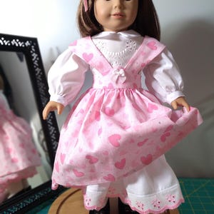 May include: A doll wearing a pink dress with white hearts and a white blouse with a lace collar. The doll has brown hair and brown eyes. The doll is wearing black shoes.