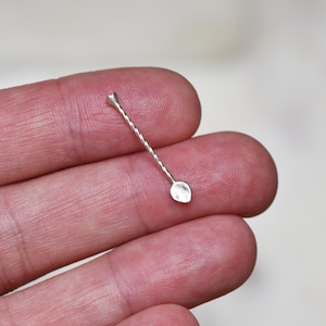 May include: A small, silver bar spoon with a twisted handle and a small spoon-shaped end. The spoon is held in a person's hand, showcasing its delicate design. This barware accessory is ideal for mixing cocktails.