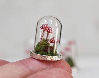 May include: Miniature glass cloche with two red and white spotted mushrooms and green moss. The cloche has a silver-colored base. The mushrooms are inside a clear glass dome, creating a whimsical, fairy-tale scene.