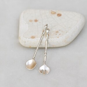 May include: Two silver-toned serving spoons, one with a spade-shaped head and the other with a fork-shaped head, rest on a smooth, off-white stone. The spoons have long, slender handles and are likely used for serving food.