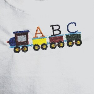 May include: White t-shirt with an embroidered train design. The train cars are yellow, red, and green, with the letters "A", "B", and "C" embroidered above them. The train engine is blue and brown.