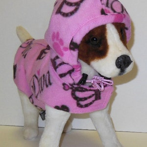 May include: A pink fleece dog hoodie with a brown and pink print of crowns, hearts, and paw prints. The hoodie has a hood and a zipper closure. The text "Diva" is printed on the front of the hoodie.