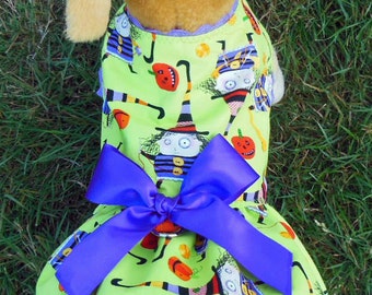 May include: A green and purple dog dress with a witch and pumpkin print. The dress has a purple satin bow and a lace trim.