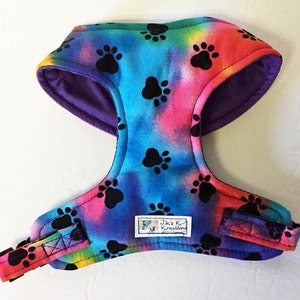 May include: A colorful dog harness with a tie-dye pattern and black paw prints. The harness is purple on the inside and has a black adjustable strap. The harness has a label that says "Jin's Kreations".