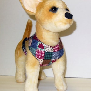 May include: A plush toy chihuahua dog with tan fur, black nose, and dark eyes. The dog wears a patchwork vest with red hearts and blue, green, and burgundy squares. The toy is standing on a white surface.