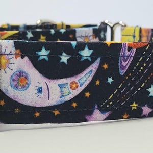 May include: A black dog collar with a celestial design. The fabric features a pattern of stars, planets, and a crescent moon in shades of yellow, blue, purple, and white. The collar has a silver metal buckle.