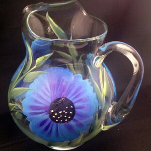 May include: A clear glass pitcher with a blue and purple flower painted on the front. The flower has a dark blue centre with yellow dots.