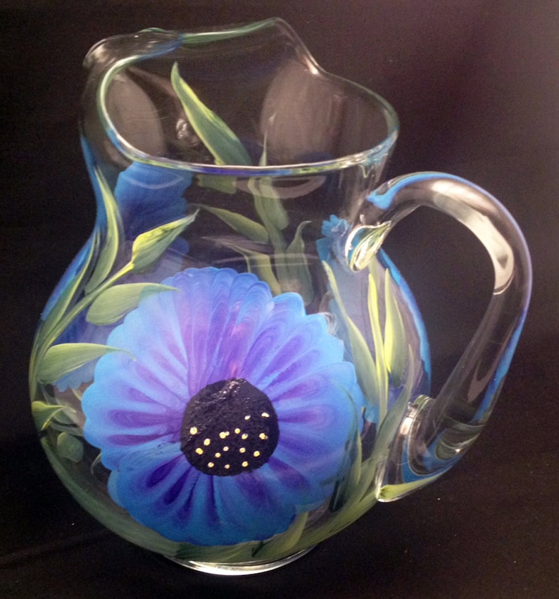 May include: A clear glass pitcher with a blue and purple flower painted on the front. The flower has a dark blue centre with yellow dots.