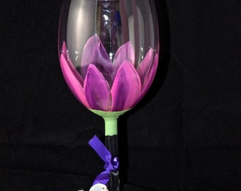 May include: A hand-painted wine glass featuring a pink flower design. The glass has a green stem and a purple ribbon tied around it. A floral patterned tag is attached to the ribbon.