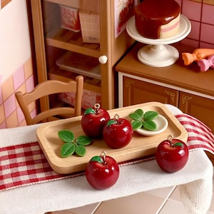 Può includere: Quattro ciondoli in miniatura a forma di mela rossa con piccole foglie verdi e anelli metallici dorati sulla parte superiore. Le mele sono disposte su un vassoio rettangolare in legno chiaro, accompagnate da due piccoli set di foglie verdi.
