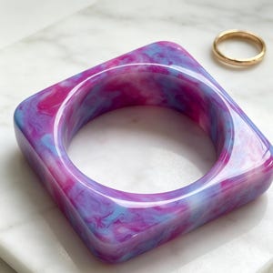 May include: A square-shaped bangle bracelet featuring a marbled pattern of vibrant pink, purple, and light blue swirls. The bangle has a smooth, glossy finish and a rounded interior opening. A thin, plain gold-toned metal ring sits nearby.