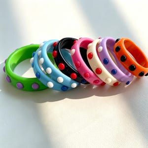 May include: A collection of seven circular bangle bracelets made of smooth, glossy resin. Each bracelet features a distinct solid color, including green, light blue, black, pink, cream, lavender, and orange, accented with small, raised dots.