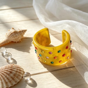 May include: A wide cuff bracelet made of yellow marbled resin, featuring a polished finish and embedded with small, round, multi-colored rhinestones in shades of red, blue, green, pink, and yellow scattered across the outer surface.
