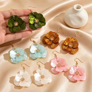 以下が含まれることがあります： 緑、青、オレンジ、ピンク、白など、さまざまな色の花の形をしたぶら下がりイヤリングのコレクション。各イヤリングには、金色のフックと小さな金色のセンターが付いています。イヤリングはサテンの表面に表示されています。