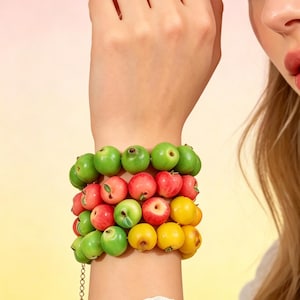 Può includere: Un bracciale a più fili caratterizzato da numerose piccole perline sferiche a forma di frutto nei toni del verde brillante, rosso e giallo. Le perline sono disposte in file, imitando l'aspetto di mele in miniatura, e sono unite da una catena.