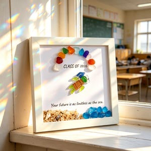 Può includere: Un'opera d'arte incorniciata con un bordo bianco, con vetro marino colorato disposto ad arco arcobaleno sopra un disegno di tartaruga marina. Il testo "CLASS OF 2026" è sopra la tartaruga e la frase "Your future is as limitless as the sea" è sotto.
