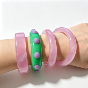 May include: A set of four bangles consisting of three translucent light pink bracelets with a smooth finish and one solid green bracelet featuring raised, rounded purple accents arranged in a single row around the outer circumference.