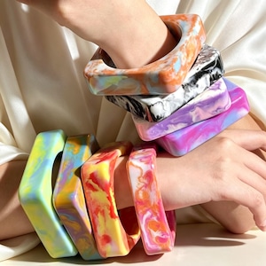 May include: A collection of square-shaped bangles featuring a marbled resin finish in various colors including orange, black, white, purple, pink, yellow, and blue. These thick, chunky bracelets have a smooth, glossy surface and rounded edges.