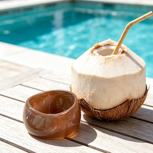 Puede incluir: Una pulsera rígida de resina marrón translúcida con una forma redondeada y orgánica, colocada junto a un coco fresco abierto con una cáscara fibrosa de color marrón claro y una pajita de bambú insertada en el agua de coco blanca.