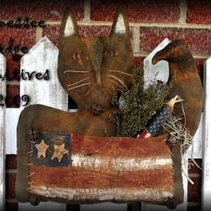May include: A rustic, brown, fabric cat figurine holding a small American flag. The cat is sitting in front of a white picket fence with the text "Tennessee Ridge Primitives ©2009" on the fence.