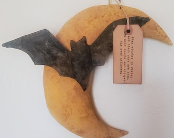 May include: A crescent moon wall decoration in a mottled yellow finish with a dark, weathered bat figure attached to the front. A rectangular tan tag hangs from the top by a string, printed with the text: When witches go riding, and black cats are seen, the moon laughs and whispers, tis near Halloween.