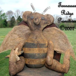 May include: A rustic, handmade bee-shaped plush doll with brown and yellow stripes, burlap wings, and wire antennae. The doll has button eyes and is sitting on a wooden surface. The text "Tennessee Ridge Primitives ©2007" is visible.