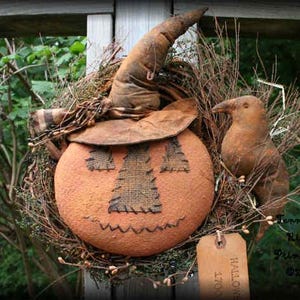 Puede incluir: Una rústica corona de Halloween con una calabaza naranja con una cara cosida, un sombrero de bruja y un pájaro de tela. La corona está hecha de ramitas y vegetación, con una etiqueta que dice "Halloween". El sombrero es marrón.