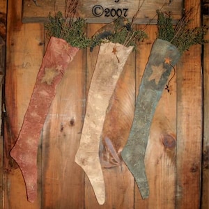 May include: Three rustic Christmas stockings in red, beige, and blue-green hang on a wooden door. Each stocking has a star and is filled with greenery. The text "Tennessee Ridge Primitives ©2007" is above the stockings.