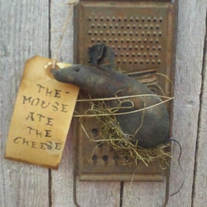 May include: A rustic, handmade mouse figurine made of fabric and wire, with a brown and black color scheme. The mouse is positioned on a vintage metal grater with a brown patina. A small tag with the text "THE MOUSE ATE THE CHEESE" is attached to the mouse.