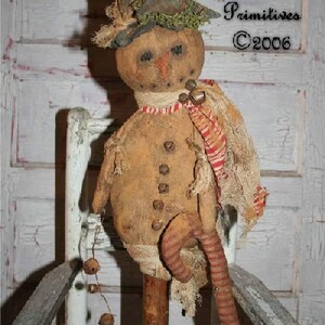 May include: A rustic snowman doll with a tall, weathered hat and a scarf with red stripes. The doll has button details and dangling bells. The text "Tennessee Ridge Primitives ©2006" is visible in the background. The snowman is seated on a distressed wooden chair.