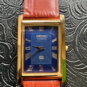 May include: A gold-toned Seiko Quartz watch with a rectangular face and a dark blue dial. The watch features gold Roman numerals and hands, and a red leather band. The watch face has the Seiko Quartz logo.