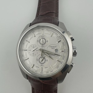 May include: Silver-toned Tissot chronograph watch with a light-colored face and a brown leather strap. The watch features multiple sub-dials, a date display, and a tachymeter scale. The strap is a dark brown color.