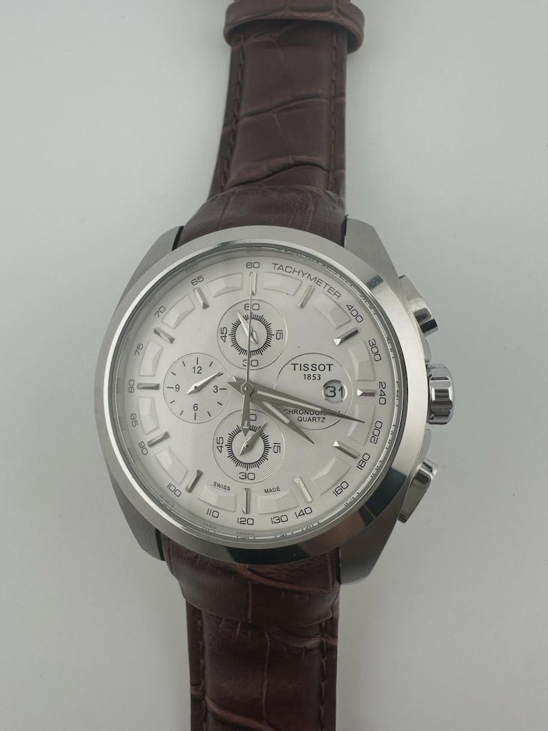 May include: Silver-toned Tissot chronograph watch with a light-colored face and a brown leather strap. The watch features multiple sub-dials, a date display, and a tachymeter scale. The strap is a dark brown color.