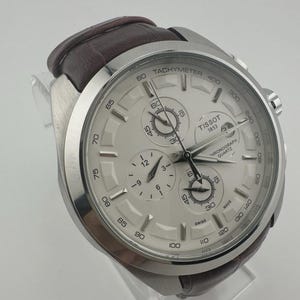 Op de afbeelding: Een zilverkleurig Tissot chronograaf horloge met een witte wijzerplaat en een bruine leren band. Het horloge heeft meerdere sub-wijzerplaten, een tachymeterschaal en de tekst "TISSOT 1853". Het horloge wordt op een transparante standaard getoond.