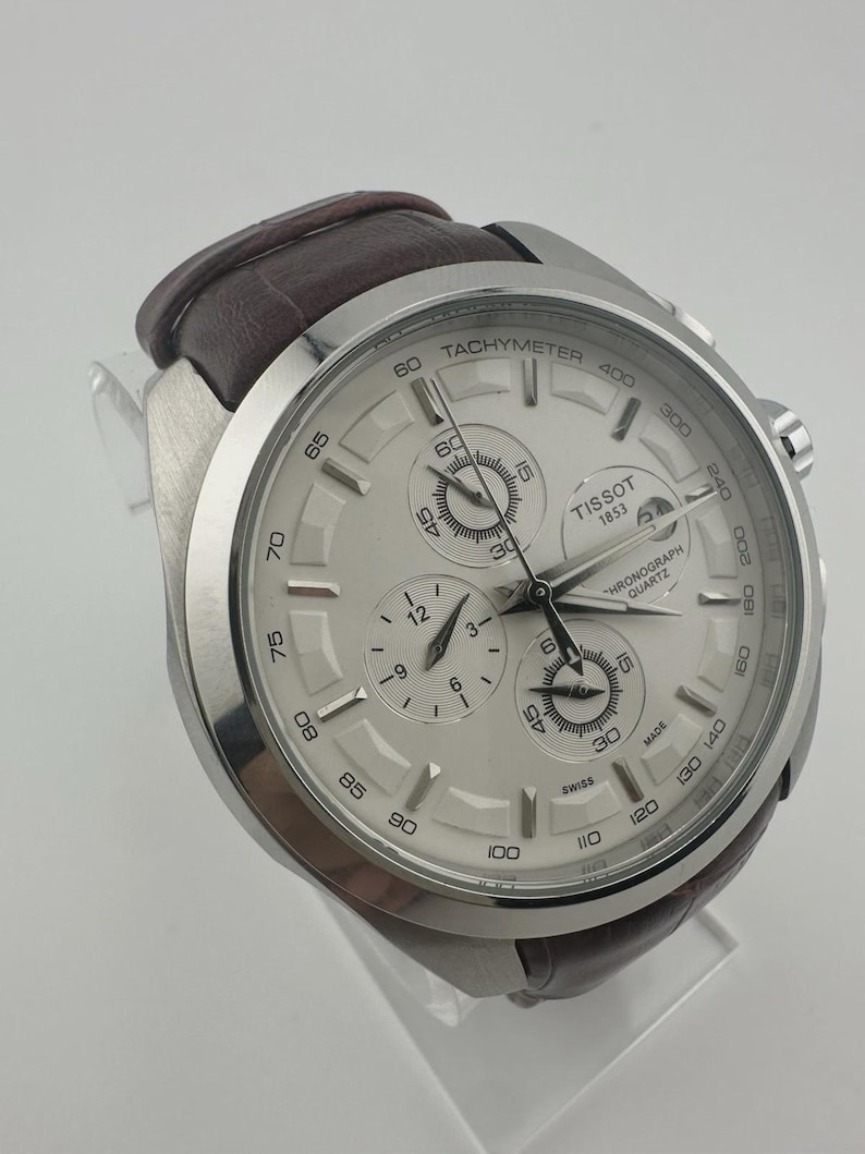 May include: A silver-toned Tissot chronograph watch with a white face and a brown leather strap. The watch features multiple sub-dials, a tachymeter scale, and the text "TISSOT 1853". The watch is displayed on a clear stand.