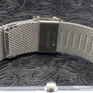 May include: Close-up of a silver stainless steel watch band with a mesh design. The band is secured with a clasp that reads "STAINLESS STEEL". The watch band is on a black textured surface.