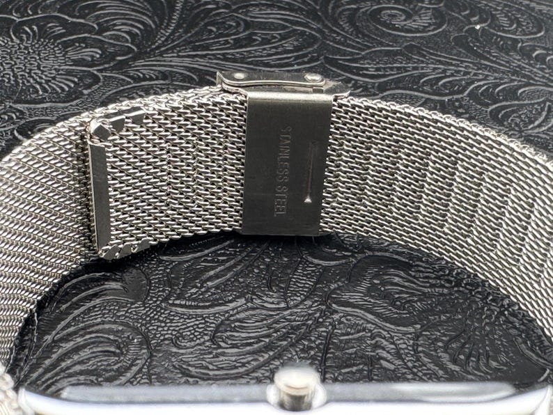 May include: Close-up of a silver stainless steel watch band with a mesh design. The band is secured with a clasp that reads "STAINLESS STEEL". The watch band is on a black textured surface.