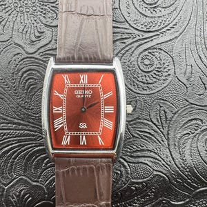 May include: A rectangular Seiko Quartz watch with a red face and silver casing. The watch features white Roman numerals and a brown leather band. The watch face displays the Seiko Quartz logo. The watch is a classic timepiece.