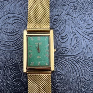 May include: A gold-toned Seiko Quartz watch with a rectangular green face and gold mesh band. The watch face features Roman numerals and the Seiko Quartz logo. The watch is displayed on a textured black surface.