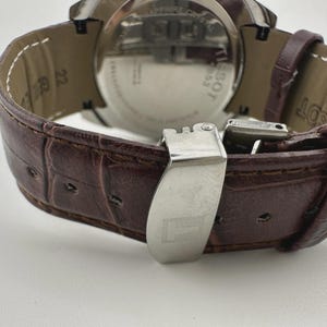 May include: Close-up of a brown leather watch strap with a stainless steel clasp. The strap has a textured, crocodile-like pattern. The watch face is visible, with the brand name "Tissot" and other details.