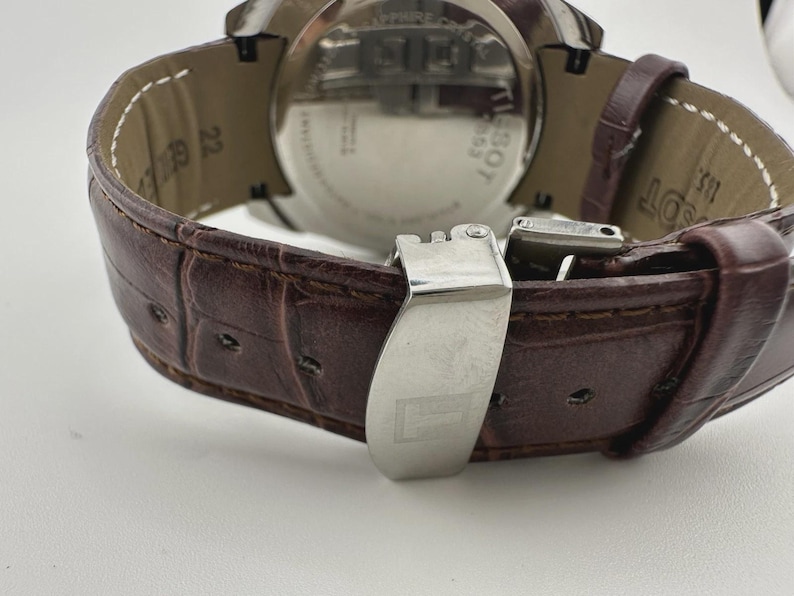 May include: Close-up of a brown leather watch strap with a stainless steel clasp. The strap has a textured, crocodile-like pattern. The watch face is visible, with the brand name "Tissot" and other details.