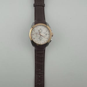 May include: A silver and gold-tone wristwatch with a white face and brown leather band. The watch features multiple dials and a date display. The band has a crocodile pattern. The watch is a luxury item.