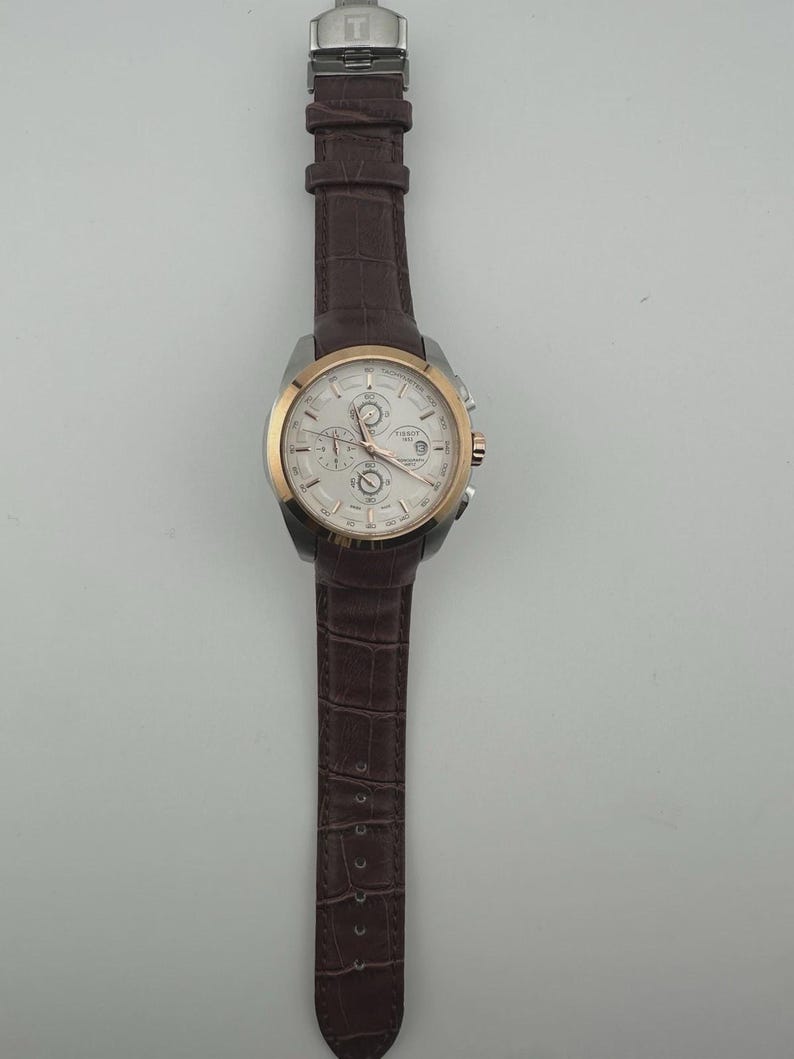 May include: A silver and gold-tone wristwatch with a white face and brown leather band. The watch features multiple dials and a date display. The band has a crocodile pattern. The watch is a luxury item.